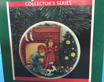 May include: A miniature porcelain plate ornament from the Collector's Series. The plate depicts two children and a dog near a Christmas tree and fireplace. Dated 1989, it includes a plate stand. The plate is a festive item.