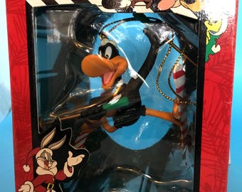 May include: A Looney Tunes Christmas ornament featuring Daffy Duck in a Santa hat, packaged in a red box with cartoon characters. The box reads "COLLECTIBLE ORNAMENTS". The ornament is black, orange, and green.