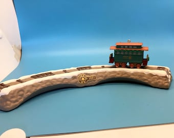 May include: A decorative train ornament featuring a green and orange caboose on a snow-covered track. The track is curved and has a gold emblem with the text "Club & Co. R.R. 1991". The ornament is set against a blue background.