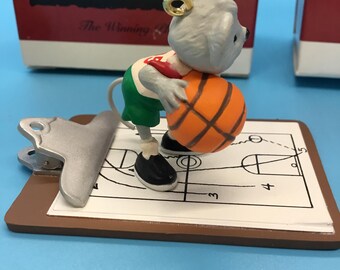 May include: A figurine of a grey mouse in a red and green basketball uniform, holding an orange basketball. The mouse stands on a clipboard with a basketball court diagram. The figurine has a gold hoop earring.