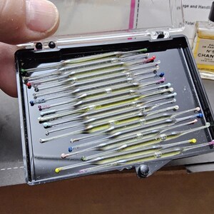 May include: A black plastic case containing numerous glass capillary tubes with colored tips. A small bottle of Chanel No. 5 perfume is visible in the background. The case is held by a person's hand.