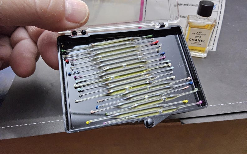 May include: A black plastic case containing numerous glass capillary tubes with colored tips. A small bottle of Chanel No. 5 perfume is visible in the background. The case is held by a person's hand.