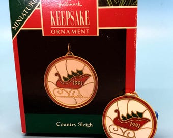 May include: A 1991 Hallmark Keepsake Ornament with a red sleigh design and green accents on a white background. The round ornament has a gold border and is presented in a decorative box. The box reads "Country Sleigh".