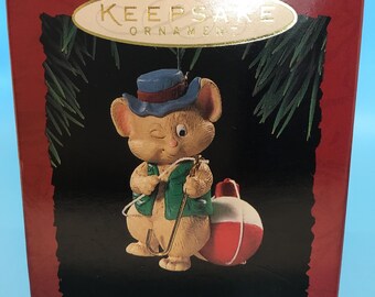 May include: A Keepsake ornament depicting a cartoon mouse wearing a blue hat and green vest, holding a fishing rod. The ornament includes a red and white bobber and the text "Merry Fishmas". The background is dark with green accents.