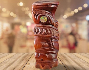 May include: A carved wooden tiki statue with a polished, reddish-brown finish. The statue features intricate carvings, including facial details and geometric patterns. A circular, iridescent inlay is set into the face. The tiki is displayed on a wooden surface.