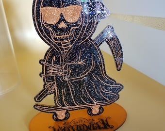 May include: A Halloween decoration depicting the Grim Reaper wearing sunglasses and holding a scythe, riding a skateboard. The figure is black with glitter and copper-coloured details. The orange base reads "Happy Halloween".