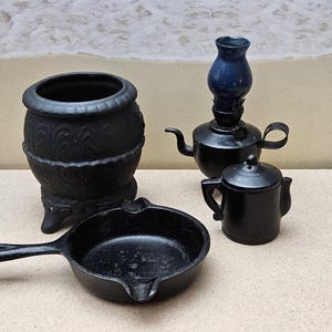 May include: A collection of miniature black cast iron kitchenware and decorative items. Includes a small frying pan, a pot, a teapot, a small pitcher, and an oil lamp with a blue glass top. The items are arranged on a light brown surface.