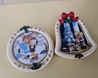 May include: Two Christmas ornaments. One is round with a child in winter clothing, the other is bell-shaped with a Christmas tree and children. Both have a cream border with holly accents and are decorative.