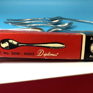 May include: A red box of Diplomat silverware with a spoon illustration. The box has the text "PAT. No. 33191 - 33242" and "Austria" printed on it. Several silver spoons are visible in the background.