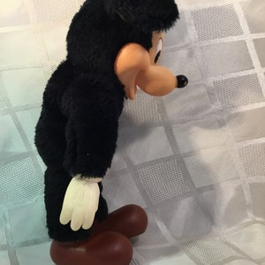 Puede incluir: Un juguete de peluche vintage de un personaje de dibujos animados, con cuerpo negro, cara color canela y pies grandes marrones. El juguete tiene guantes blancos y una cabeza negra con orejas ca&iacute;das. El juguete est&aacute; sobre una superficie blanca.