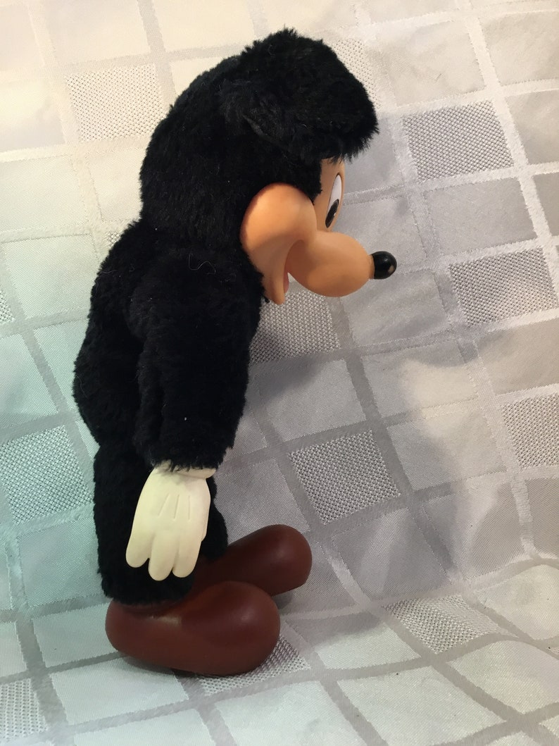 Puede incluir: Un juguete de peluche vintage de un personaje de dibujos animados, con cuerpo negro, cara color canela y pies grandes marrones. El juguete tiene guantes blancos y una cabeza negra con orejas ca&iacute;das. El juguete est&aacute; sobre una superficie blanca.