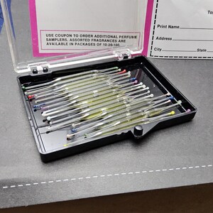 May include: A black plastic case with a clear lid containing multiple glass perfume scent strips with colored tips. A bottle of Chanel No. 5 perfume sits to the right. The case is on a dark surface. Text on the lid reads: USE COUPON TO ORDER ADDITIONAL PERFUME SAMPLERS.