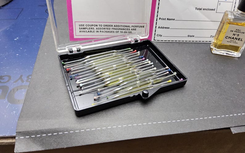 May include: A black plastic case with a clear lid containing multiple glass perfume scent strips with colored tips. A bottle of Chanel No. 5 perfume sits to the right. The case is on a dark surface. Text on the lid reads: USE COUPON TO ORDER ADDITIONAL PERFUME SAMPLERS.