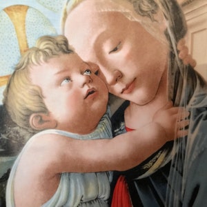 May include: A detail from a Renaissance painting depicting a mother and child. The mother, with a serene expression, cradles the child, who gazes upward. The artwork features soft colors and delicate details, with a halo visible in the background.