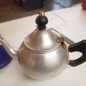 May include: A small, silver-colored teapot with a black handle and a decorative black and gold knob on the lid. The teapot has a spout and a hinged lid with a metal clasp. The teapot is sitting on a white surface.