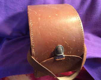 May include: A brown leather camera bag with a curved top and a strap. The bag features a metal clasp and shows signs of wear, with scratches on the surface. The stitching is visible along the edges.