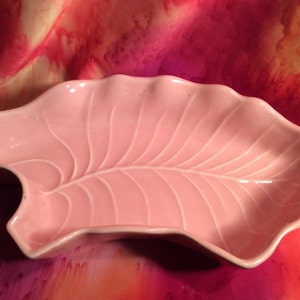 May include: A light pink ceramic leaf-shaped dish with a white rim. The dish has detailed veining and a wavy edge. This decorative dish is ideal for serving snacks or as a decorative accent.