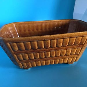 May include: A rectangular brown ceramic planter with a textured, ridged design. The planter has a wide opening and sits on small feet. The exterior features a repeating pattern of vertical, rounded ridges. The planter is suitable for indoor or outdoor use.