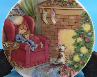 May include: A decorative plate depicting a Christmas scene. Two children are asleep in a red armchair near a fireplace with stockings. A dog sits near a Christmas tree with presents. The plate has a blue background.