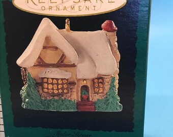 May include: A Keepsake Ornament of a Tudor House from the Old English Village Collector's Series. The miniature house has a snow-covered roof, a red chimney, and green shrubbery. The box is dark green with gold lettering.
