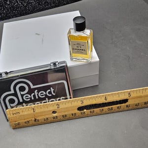 May include: A small bottle of Chanel No. 5 perfume, a black and clear case with the text "Perfect Retreaders", a white box, and a wooden ruler with inch measurements. The ruler is approximately 12 inches long.
