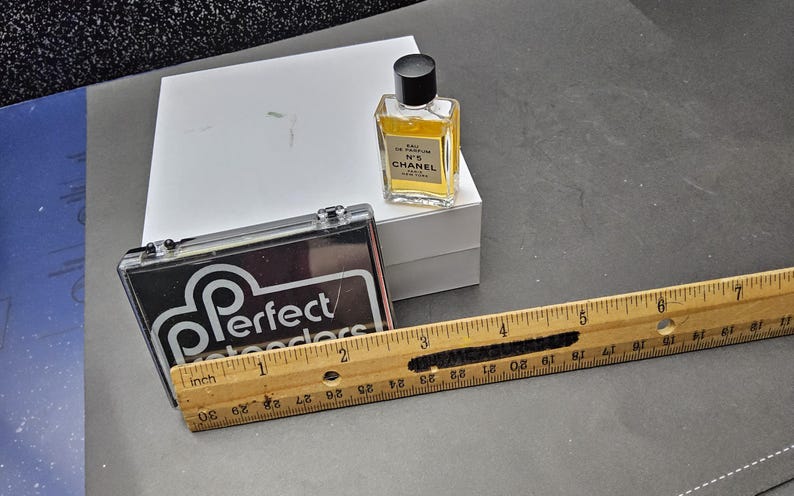 May include: A small bottle of Chanel No. 5 perfume, a black and clear case with the text "Perfect Retreaders", a white box, and a wooden ruler with inch measurements. The ruler is approximately 12 inches long.
