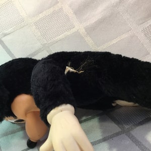 Puede incluir: Un juguete de peluche negro con cara y manos color crema. El juguete est&aacute; tumbado sobre una superficie blanca con un patr&oacute;n de diamantes. El brazo del juguete est&aacute; extendido y la cara est&aacute; girada hacia el espectador.