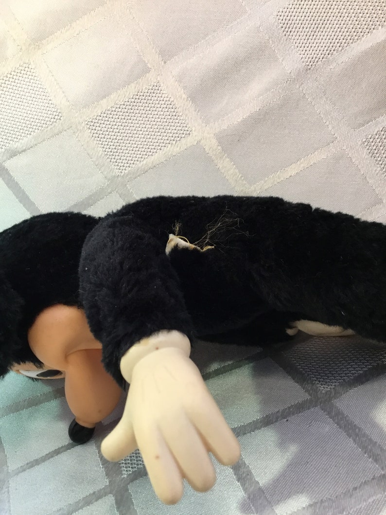 Puede incluir: Un juguete de peluche negro con cara y manos color crema. El juguete est&aacute; tumbado sobre una superficie blanca con un patr&oacute;n de diamantes. El brazo del juguete est&aacute; extendido y la cara est&aacute; girada hacia el espectador.