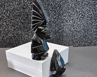 May include: A black carved sculpture of a figure with a tall, fan-shaped headdress. The sculpture is on a white box, with a detached piece nearby. The background is a speckled black and white pattern.
