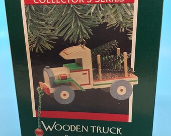 May include: A boxed Christmas ornament depicting a wooden truck hauling a Christmas tree. The truck is cream, green, and red, with grey wheels. The box is green with red accents and the text "COLLECTOR'S SERIES" and "Wooden Truck Dated 1989".
