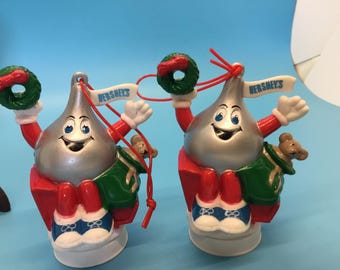 May include: Two Hershey's Kiss ornaments with smiling faces, holding wreaths and banners that read "Hershey's". The ornaments have red arms and legs, green jackets, and blue and white shoes. A small teddy bear is on the green jacket.