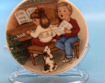 May include: A decorative plate depicting a Christmas scene. Two children are singing and playing the piano, with a small dog in front. The plate has a glossy finish and is displayed on a clear plastic stand.