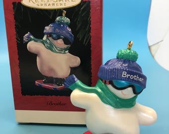 May include: A snowman ornament on a red snowboard, wearing a blue hat with the word "Brother", a green scarf, and sunglasses. The ornament is next to a red box with the words "Keepsake Ornament".