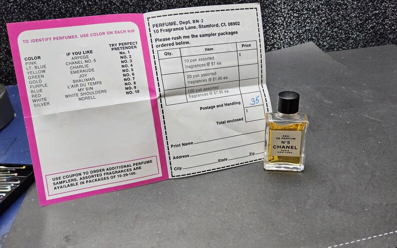 May include: A small bottle of Chanel No. 5 Eau de Parfum sits next to a pink and white perfume identification chart and an order form. The perfume bottle has a black cap and a gold label. The order form lists sampler packages.