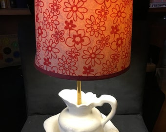 May include: A table lamp featuring a white ceramic pitcher and basin base with a gold-toned central rod. The cylindrical lampshade is made of a translucent material with a repeating red floral pattern and a solid red trim along the bottom edge.