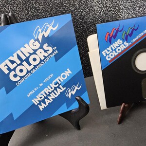 May include: Two vintage computer software items. One is a blue instruction manual with the text "FLYING COLORS COMPUTER GRAPHICS SOFTWARE". The other is a black floppy disk with the same logo and text. Both are displayed on small stands.