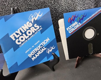 May include: Two vintage computer software items. One is a blue instruction manual with the text "FLYING COLORS COMPUTER GRAPHICS SOFTWARE". The other is a black floppy disk with the same logo and text. Both are displayed on small stands.