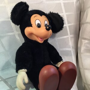 Puede incluir: Un mu&ntilde;eco de peluche vintage de Mickey Mouse. El mu&ntilde;eco tiene un cuerpo negro, orejas grandes negras, una cara bronceada y zapatos marrones grandes. El mu&ntilde;eco tiene guantes blancos y est&aacute; sentado sobre una superficie blanca estampada.