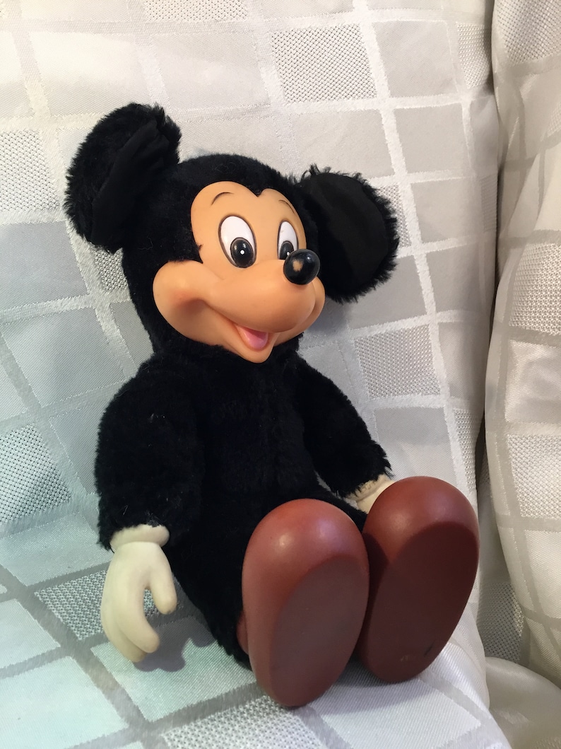 Puede incluir: Un mu&ntilde;eco de peluche vintage de Mickey Mouse. El mu&ntilde;eco tiene un cuerpo negro, orejas grandes negras, una cara bronceada y zapatos marrones grandes. El mu&ntilde;eco tiene guantes blancos y est&aacute; sentado sobre una superficie blanca estampada.
