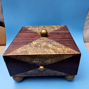 May include: A rectangular wooden box with a dark brown finish and gold accents. The lid and sides feature a geometric design with gold paint. The box has a gold knob on the top and bottom, and rests on four gold ball feet.