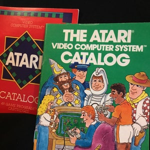May include: Two vintage Atari video game catalogs. One is red with a blue and green geometric design and the word "ATARI" in white. The other is green and features cartoon characters and the text "THE ATARI VIDEO COMPUTER SYSTEM CATALOG".