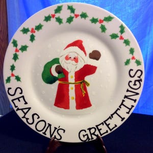 May include: A white ceramic plate with a hand-painted Santa Claus design. The plate features the text "SEASON'S GREETINGS" in black, with holly and berry accents. Santa is wearing a red suit and holding a green sack.