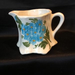 May include: A small, white ceramic creamer with a handle and a spout. The creamer is decorated with a cluster of blue flowers with yellow centres and green leaves. The creamer is on a black surface.