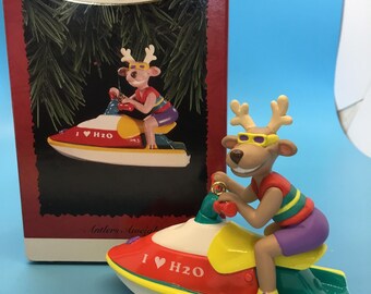 May include: A Christmas ornament depicting a cartoon reindeer in sunglasses on a red, yellow, and green jet ski. The jet ski has the text "I ♥ H2O". The ornament is in its original box.