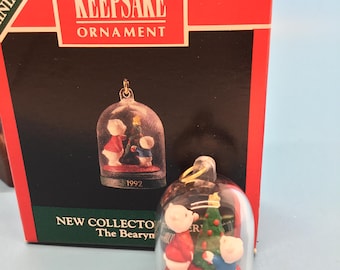May include: A 1992 Hallmark Keepsake Ornament. The ornament features two bear figurines and a Christmas tree inside a clear dome. The base is red, and it has a gold-coloured hanger. The box reads "Keepsake Ornament".