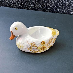 May include: A white ceramic duck-shaped dish with a yellow beak and floral accents. The dish has a small bowl-like indentation on its back. The duck's wings and body are decorated with yellow floral patterns.