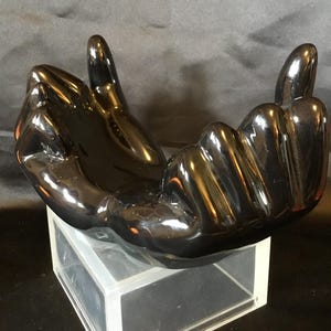 May include: A glossy black sculpture of two hands forming a gesture. The hands are positioned to create a bowl-like shape, with the fingers pointing upwards. The sculpture is placed on a clear, rectangular base.