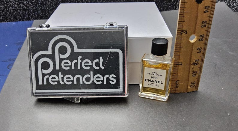 May include: A small bottle of Chanel No. 5 perfume with a black cap, next to a clear plastic case with the words "Perfect Pretenders" in white on a black background. A wooden ruler is also present.
