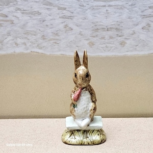 May include: A ceramic figurine of a rabbit holding a carrot. The rabbit is brown and white, with long ears and a seated pose. It is sitting on a white block with a grass-like base. The background is a blurred beach scene.