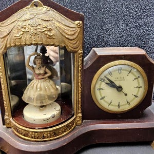 May include: Vintage wooden clock with a ballerina figurine inside a decorative frame. The clock features a gold-toned face with black numerals and hands. The ballerina is wearing a pink and beige dress.